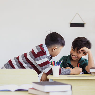 Two children interacting at a study table with books and colored pencils, one child smiling while writing and the other leaning in with interest.