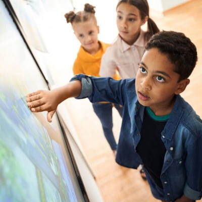 Primary aged students gathered around, and looking up at an interactive display. In the center of the image, a young boy looking amazed at the content on the screen.