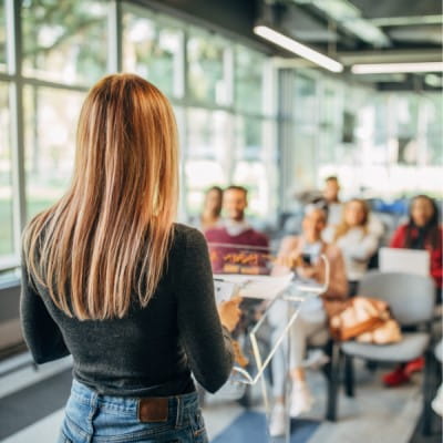 A female professional standing at a podium and presenting to a room of people.