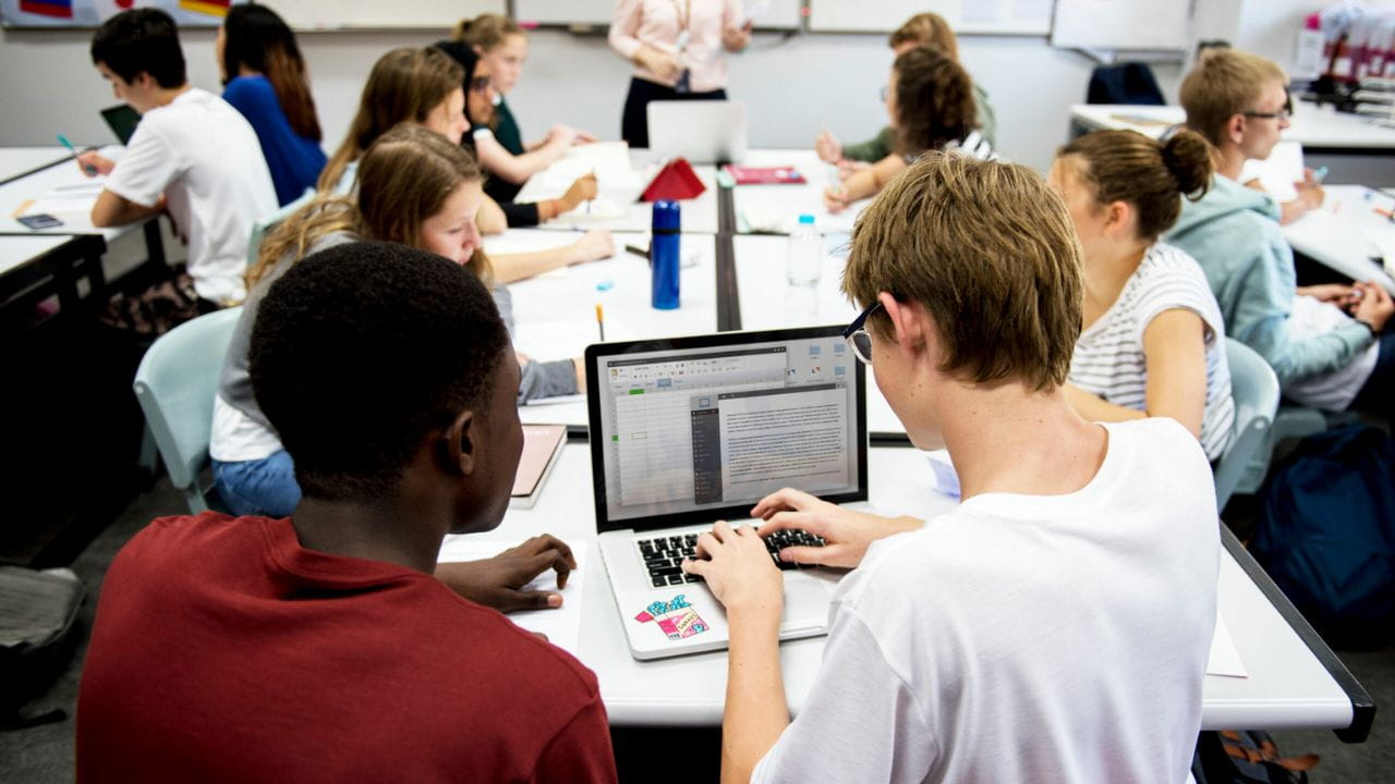 A classroom full of high school students, with a view over the shoulders of two students engaged in group work.