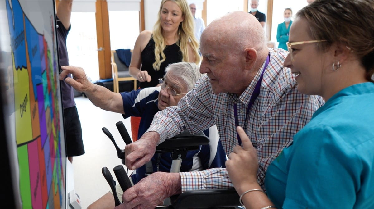 Elderly residents in a care home engaging with colorful activities on a SMART Board, assisted by a smiling facilitator.