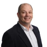 in black blazer and patterned shirt smiling against a white background.