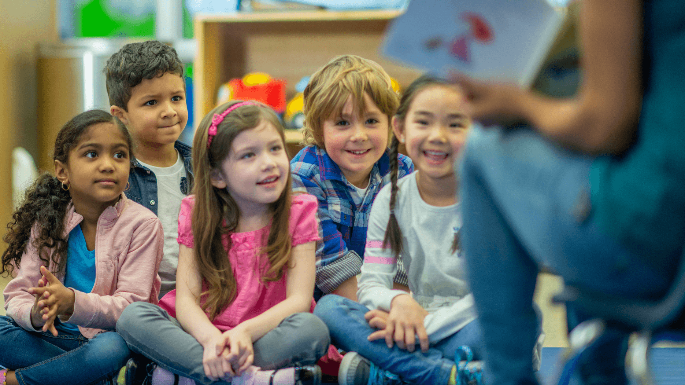 5 Kindergarten students sitting and listening to their teacher who is out of focus.